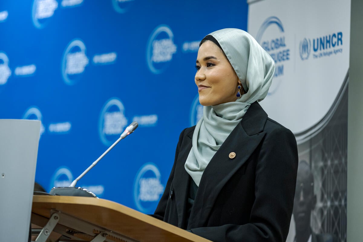 Una mujer, que lleva puesto un hijab, pronuncia un discurso desde un podio. Frente a ella, un delgado micrófono; detrás, un fondo azul con el logotipo del Foro Mundial sobre los Refugiados.,