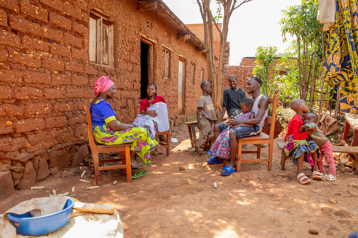 Un grupo de personas sentadas a la sombra afuera de una casa hecha de adobe